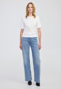 Weißes Kurzarmoberteil mit goldener quadratischer Schnalle, kombiniert mit hellblauen weit geschnittenen Jeans. Schwarze Knöchelschuhe runden das Outfit ab.