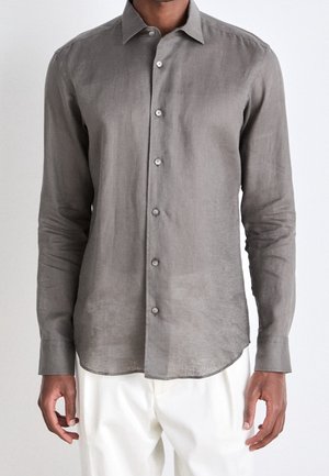 Homme portant une chemise grise à manches longues boutonnée avec un col et un pantalon blanc, debout devant un fond clair uni.