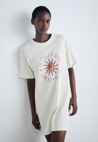Oversized cream cotton t-shirt dress with short sleeves, featuring a sun graphic and text reading "SLOW DOWN" in warm tones.