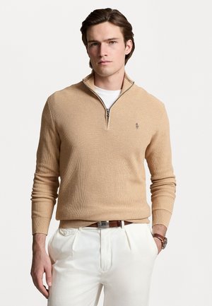 Pull en tricot beige avec col zippé, poignets côtelés et petit logo sur la poitrine, porté sur une chemise blanche et assorti à un pantalon blanc.