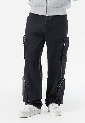 Pantalones cargo negros con múltiples bolsillos con cremallera, combinados con zapatillas blancas y una chaqueta gris claro con cremallera.