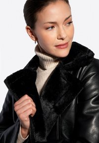 Czarna skórzana kurtka z pluszowym czarnym kołnierzem, z zamkiem błyskawicznym i teksturowanym wykończeniem. Nosi na sobie jasnoszary golf.