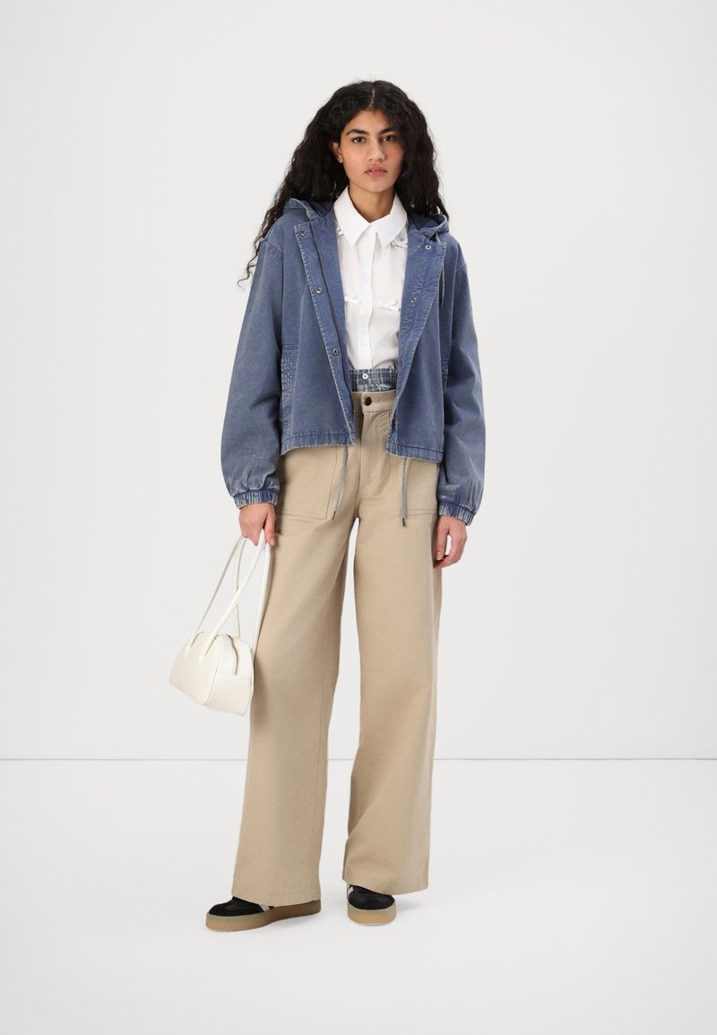 Veste en denim avec capuche, couleur bleu clair, poignets élastiques, associée à un pantalon beige à jambes larges et un petit sac à main blanc. Chaussures noires avec une semelle beige.