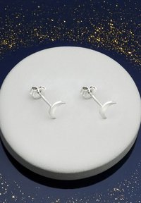 Silverhalvmåne-studörhängen med en polerad yta, monterade på en vit utställningsyta mot en mörk, glittrig bakgrund.