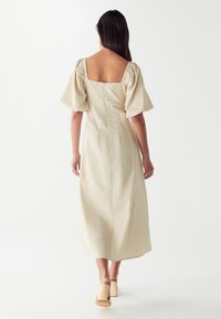 Bež haljina od lana s napuhanim rukavima, kvadratnim izrezom na leđima i dužinom do gležnjeva. Ima uski bodice i rašireni kroj.