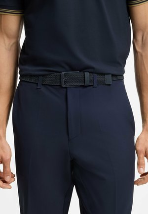 Cintura nera intrecciata con fibbia rettangolare, indossata sopra pantaloni blu navy. Il design include materiale testurizzato con dettagli di cinturino regolabile.