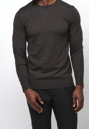 Homme portant un pull gris foncé ajusté à manches longues et un pantalon noir, debout devant un fond blanc uni.