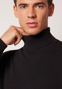 Schwarzer Rollkragenpullover aus einem weichen, strukturierten Stoff. Nahaufnahme des Kragens und des Gesichts, die Kragen Details und glatte Hauttöne zeigen.