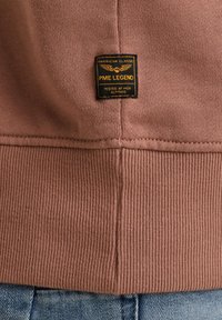 Bruine sweatshirtstof met geribbelde zoom, voorzien van een zwart label met gouden tekst en vleugels, bevestigd aan de onderrand.