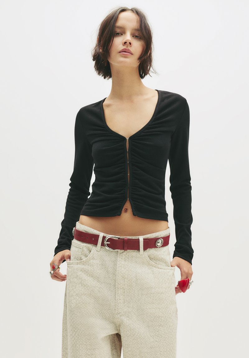 Top aderente nero a maniche lunghe con scollo rotondo e fronte arricciato. Abbinato a pantaloni di colore chiaro con un motivo a rombi e una cintura rossa.