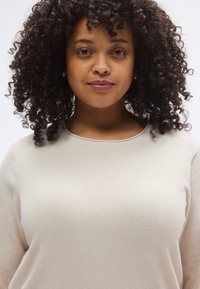 Beiger, weich strukturierter Pullover mit rundem Ausschnitt und langen Ärmeln, mit minimalen Nahtdetails. Lockiges Haar umrahmt das Gesicht.