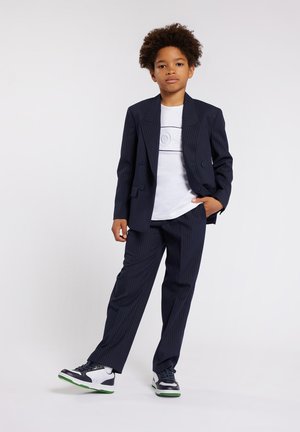 Completo a righe navy con giacca doppiopetto, abbinato a una t-shirt bianca e sneaker nere con dettagli verdi.
