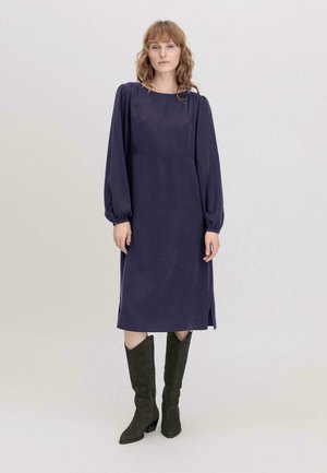 Navyblaue lange Kleid mit quadratischem Ausschnitt, gerafften Ärmel-Details, seitlichen Schlitzen und glattem Stoff, kombiniert mit knielangen grünen Stiefeln.