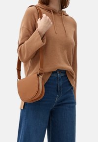 Brun läderaxelväska med en böjd klaffdesign, justerbar rem och texturerad yta. Matchad med en oversized beige tröja och blå jeans.