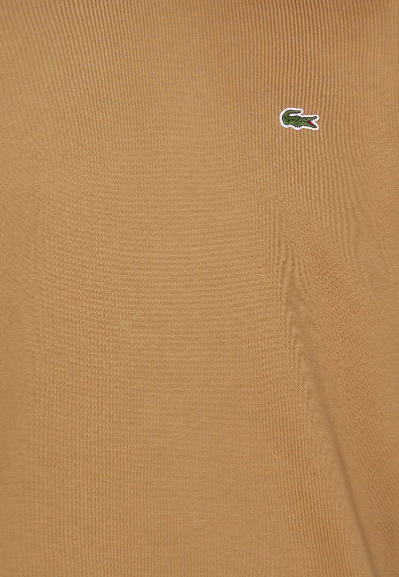 Kurzärmliges T-Shirt mit Rundhalsausschnitt in einem warmen Beigeton, aus weicher Baumwolle. Verfügt über ein kleines, gesticktes grünes Krokodil-Logo auf der Brust.