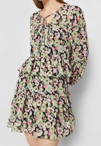 Blumenkleid mit langen Ärmeln, gerafften Schichten und einer Schleifenverschluss. Hat einen schwarzen Grund mit grünen, rosa und weißen Blumenmustern.