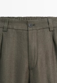 Olivgröna byxor med hög midja, elastisk midjeband, svart knappstängning, bälteshällor och subtilt fiskbensmönster.