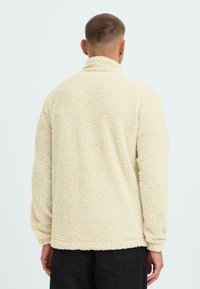 Giacca in pile color crema con una superficie morbida e testurizzata. Caratterizzata da un colletto alto e maniche lunghe. Vestibilità semplice e rilassata senza chiusure visibili.