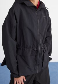 Svart zip-up jacka med huva, elastisk midja och muddar. Har en enkel design med en liten logotyp och framfickor. Lätt, strukturerad tyg.