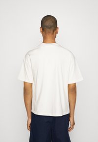 Camiseta de manga corta en color crema con cuello redondo, de corte holgado y dobladillo recto, hecha de suave tela de algodón. Se muestra la vista trasera.
