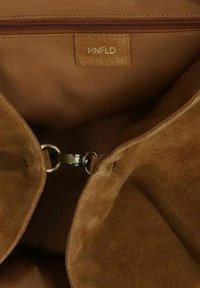 Innenraum einer braunen Wildleder-Handtasche mit einem Reißverschlussfach, goldenen Beschlägen und Markenetikett. Weiche Textur mit glatten Nahtdetails.