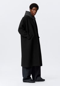 Persona joven con un abrigo largo negro sobre una sudadera con capucha gris, jeans oscuros anchos y zapatos negros texturizados, de pie contra un fondo blanco liso.