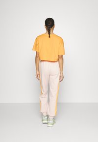 Žena stojící zády, oblečená v oranžovém zkráceném tričku, světlých růžových teplácích s oranžovými bočními pruhy a světle zelených teniskách.