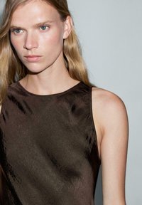 Brązowa sukienka bez rękawów o gładkiej, lekko błyszczącej teksturze. Okrągły dekolt i minimalistyczny design podkreślający delikatny połysk materiału.