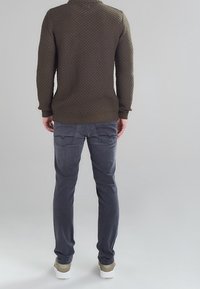 Maglione lavorato a maglia verde oliva con motivo testurizzato, abbinato a jeans grigio scuro. Le scarpe sono chiare con una suola bianca e accenti verde chiaro.