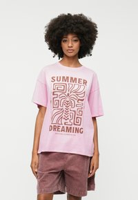 Pink oversized t-shirt featuring "SUMMER DREAMING" graphic in brown, with abstract plant and geometric designs. Worn with brown corduroy shorts.