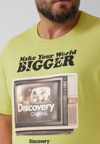 Jasnozielona koszulka z grafiką vintage telewizora z napisem "Spraw, aby Twój świat był WIĘKSZY" oraz logo "Discovery Channel".