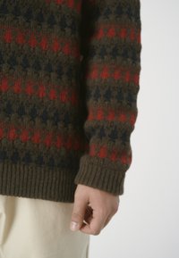 Brauner Strickpullover mit geometrischem Muster in Rot und Marineblau, mit geripptem Bündchen und strukturiertem Stoff. Eine Hand ist in der Nähe des Ärmels positioniert.