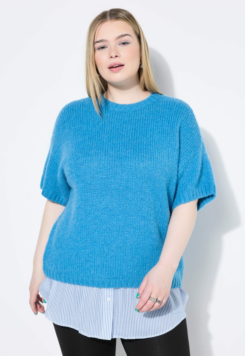 Maglione azzurro chiaro lavorato a maglia con maniche corte, indossato sopra una camicia bianca e blu a righe con orlo curvo e leggings neri.