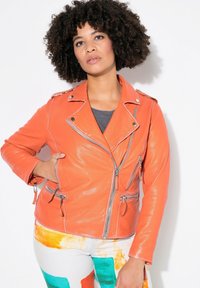 Orangene Leder-Bikerjacke mit silbernen Reißverschlüssen, ausgestattet mit Seitentaschen und einem eingeschnittenen Kragen. Kombiniert mit bunten, gemusterten Hosen.