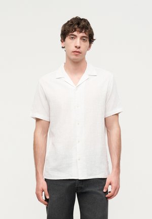 Jeune homme aux cheveux bouclés portant une chemise blanche à manches courtes boutonnée et un pantalon foncé, debout devant un fond clair uni.