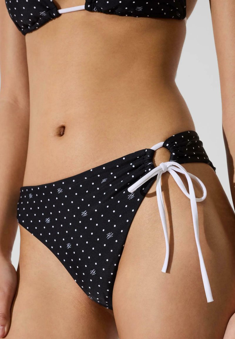 Bas de bikini noir à pois blancs et petits logos, avec un anneau latéral et un lien blanc, porté à la taille et aux hanches d'un mannequin.