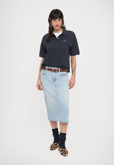 Γυναίκα με σκουροκάστανα μαλλιά φοράει μπλε ναυτικό πόλο μπλουζάκι, μίντι φούστα από ανοιχτό μπλε τζιν με καφέ ζώνη, σκούρες κάλτσες και φλατ παπούτσια με λεοπάρ σχέδιο πάνω σε λευκό φόντο.