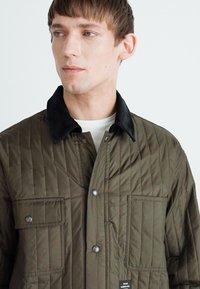 Olivgrüne gesteppte Jacke mit einem schwarzen Cordkragen, zwei Fronttaschen und metallenen Druckknöpfen, die eine strukturierte Oberfläche aufweist.