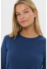 Fiatal nő hosszú szőke hajjal, kék kerek nyakú pulóvert viselve, enyhén mosolyogva egy sima fehér háttér előtt.