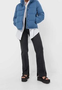 Person wearing blue puffer jacket over long white button-up shirt, black split-hem pants, and black platform sandals, standing against white background.