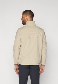 Veste légère beige avec un col montant, des poignets élastiques et un logo brodé dans le dos, associée à un pantalon sombre.