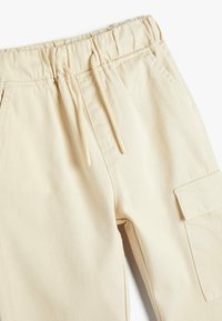 Lyse beige cargo bukser har et elastisk talje med snore, flade forlommer og en påsyet lomme på siden. Glat stoftekstur.