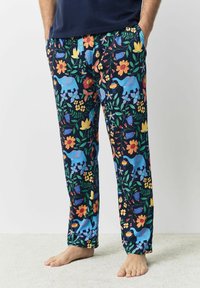 Mann in navyblauen Hosen mit bunten Blumen- und blauen Elefantenmustern, der barfuß auf einem hellen Teppich steht und die Hände in den Taschen hat.