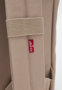 Sangle de sac à dos en tissu beige avec une surface texturée, arborant une étiquette de logo rouge Levi's cousue sur la sangle.