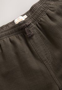 Dark olive green, textured fabric with an elastic waistband, drawstring tie, and stitched detailing. Label reads "Next Supply."