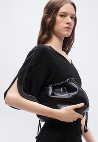 Νεαρή γυναίκα με μαύρο φόρεμα κρατώντας μια μαύρη δερμάτινη τσάντα φάκελο μπροστά σε ένα απλό λευκό φόντο.