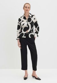 Camicia bianca e nera con grandi motivi circolari, abbinata a pantaloni neri sartoriali e mules a punta. Tessuto di texture liscia.