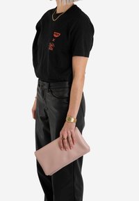 Różowa skórzana kopertówka trzymana w dłoni, wyróżniająca się gładką fakturą i subtelnym brandingiem. Czarna koszulka i skórzane spodnie dopełniają strój.