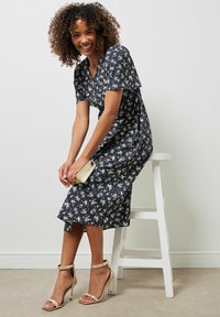 Vestido floral azul marino de tela ligera, con mangas cortas y dobladillo acampanado; combinado con tacones metálicos de tiras y un pequeño clutch dorado.