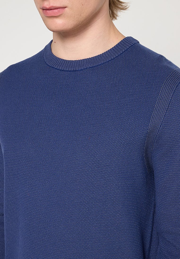 Närbild av en person som bär en strukturerad marinblå crewneck-tröja, som visar överkroppen och en del av nacken och käklinjen.